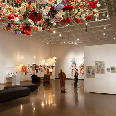A brightly-lit art gallery with white walls. Hundreds of artificial flowers hang suspended from the ceiling above the viewer, with other paintings and drawings hanging around the room.