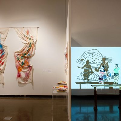 A sheet, stained with various colors of paint, hangs on a wall. To its right, Balinese shadow puppets cast an image on the back wall, against a projected white background.