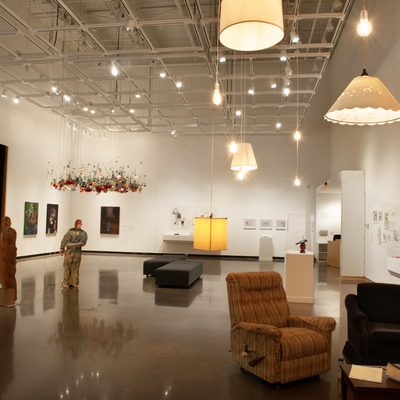 A brightly lit art gallery with white walls. The viewer stands near two arm chairs and a coffee table underneath two ceiling lamps.