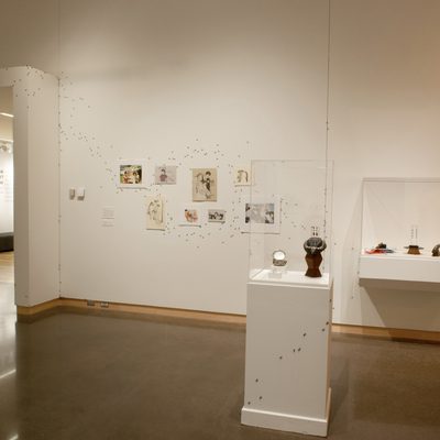 Eight drawings hang on a white wall, with jewelry displayed on pedestals to the drawings’ right. Climbing down the wall and winding around one of the pedestals are hundreds of small black ants.