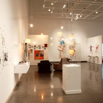 brightly-lit art gallery with white walls. On the wall to the left of the viewer, we can see several drawings mounted on the wall, with small black ants crawling around them.