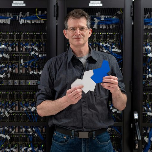 Craig S. Kaplan is a light skinned man with salt-and-pepper hair and glasses. He wears a dark gray button-up shirt and dark blue jeans. He stands in front of a wall of computer servers. He holds two interconnecting aperiodic tiles, one light gray and one bright blue.