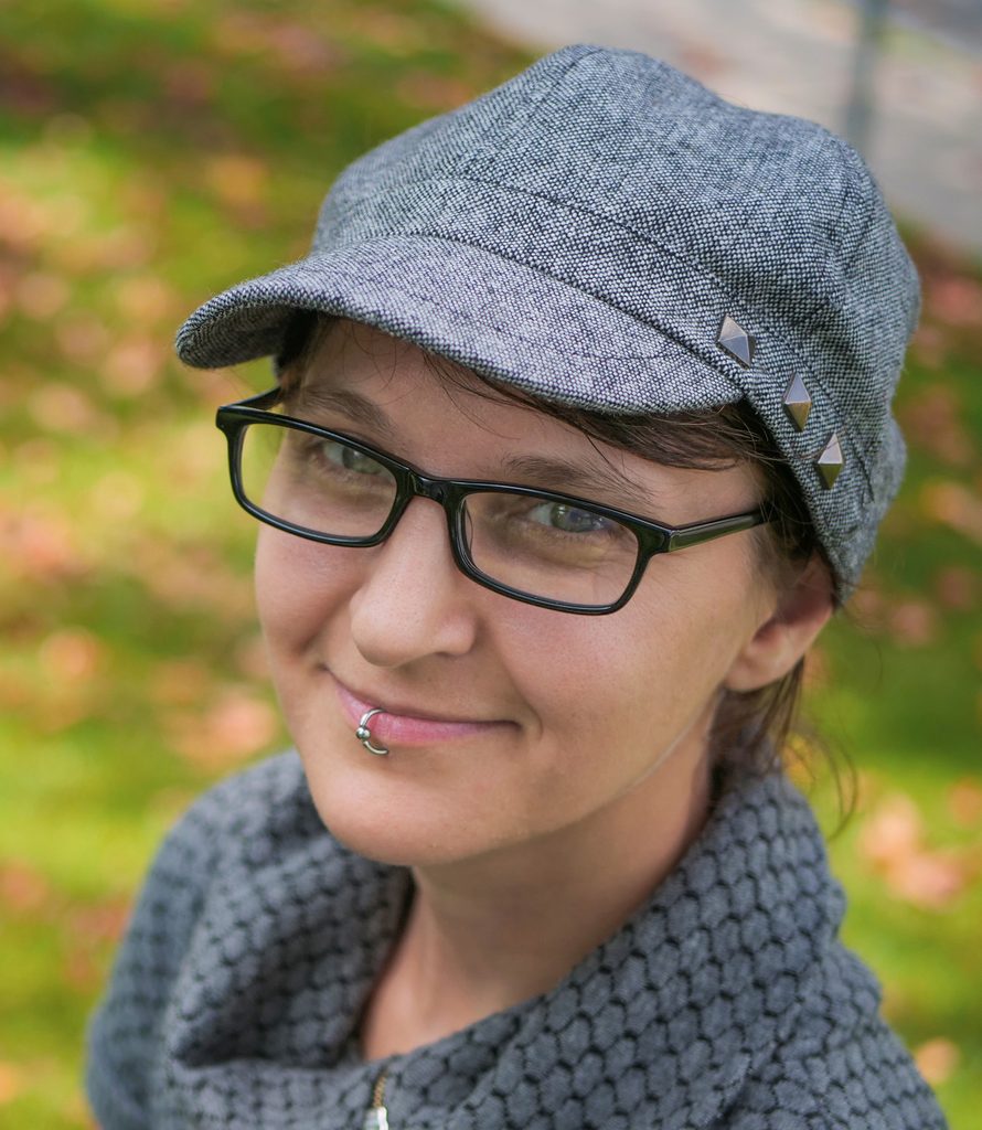 Gillian Turnham is a light-skinned woman in a medium gray brimmed cap and a gray coat with rounded collar. She has short brown hair and glasses. She wears a silver lip ring in the center of her bottom lip. She is smiling, and has a friendly look on her face.
