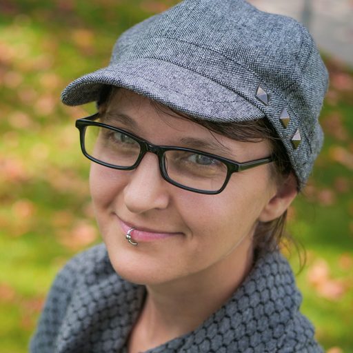 Gillian Turnham is a light-skinned woman in a medium gray brimmed cap and a gray coat with rounded collar. She has short brown hair and glasses. She wears a silver lip ring in the center of her bottom lip. She is smiling, and has a friendly look on her face.