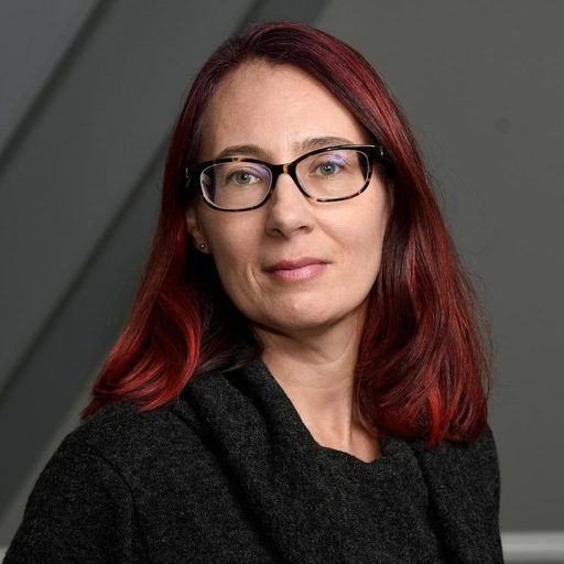 Rachel Seligman is a light skinned woman. She wears glasses and a black shirt. She has red, shoulder length hair. Behind her is a gray wall.
