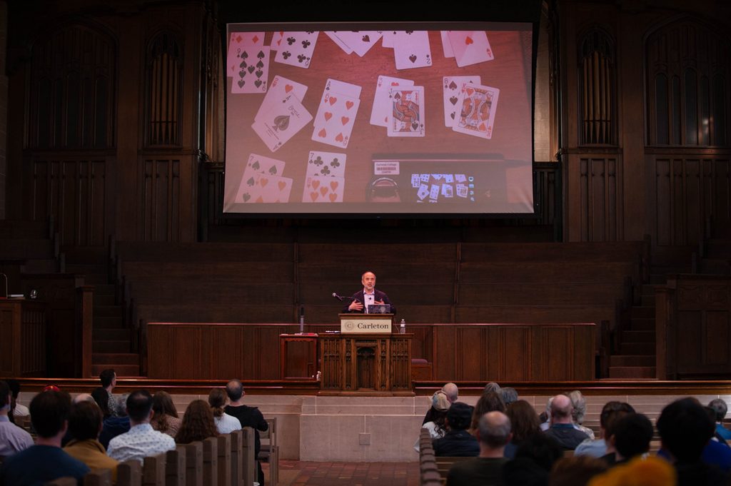 A man speaks in front of a crowd in a chapel. Above him is a large screen that has a view of his hands showing attendees a card trick.