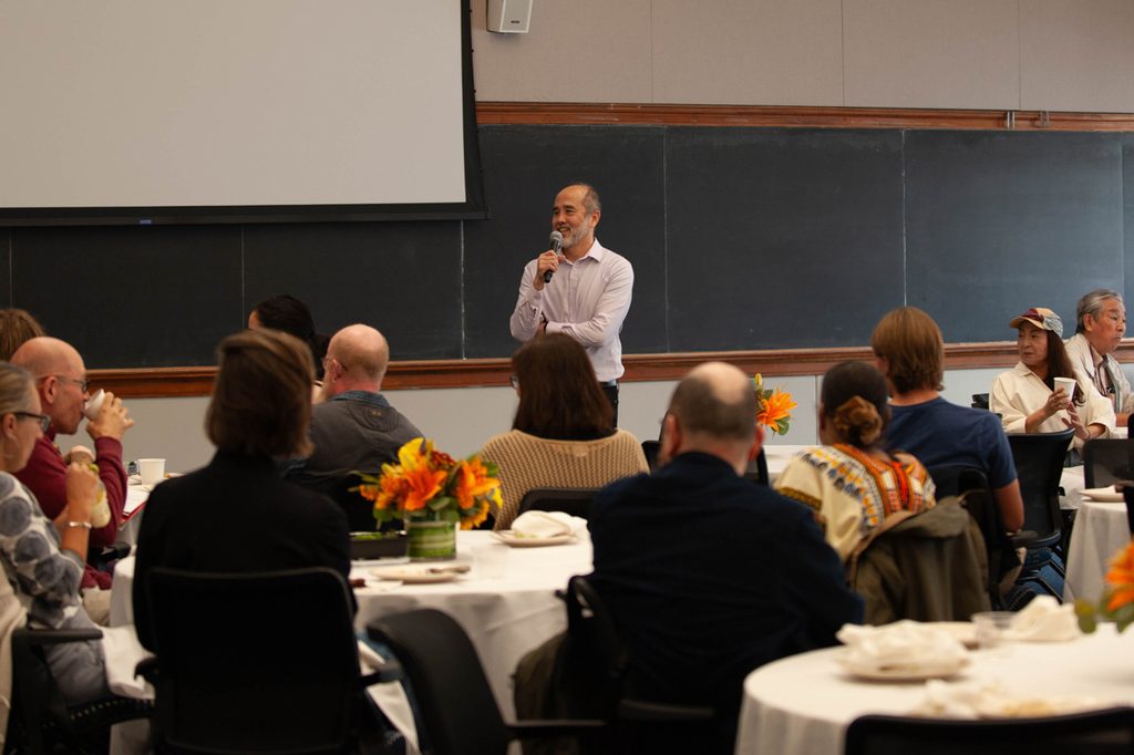 In a meeting room with chalkboards in the background, a man smiles and speaks into a microphone, seated people eat and drink.