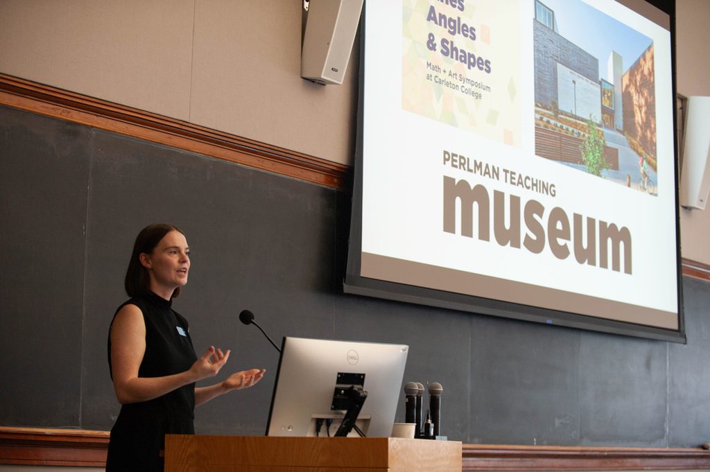 A woman speaks at a podium in a classroom with a chalkboard background. A bright screen with her presentation is in the background.
