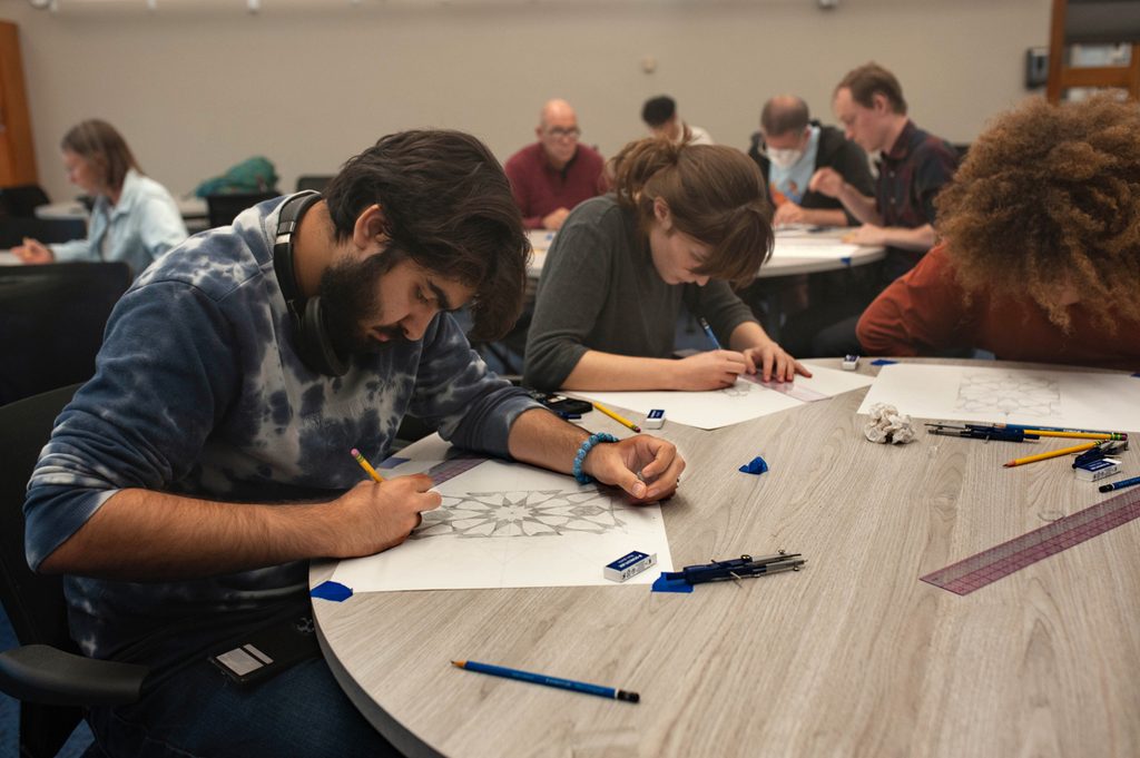 Students draw a geometric pattern at a round table. On the desk are fullers, pencils, and compasses.
