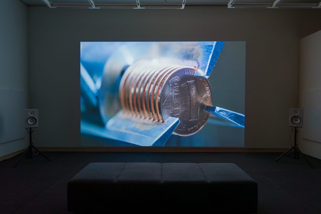 Alt text: Along the wall of a brightly-lit gallery sits a pedestal with a small vintage tv, projecting a man sitting in front of an American flag. On the wall hangs a frame artwork with a dark gray background and mounted rows of yellow checks and white labels.