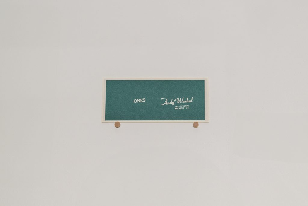 A dollar bill sized piece of paper sits on a white background. The paper is green, with a tan border and reads “ones,” and features Andy Warhol’s stamped signature.
