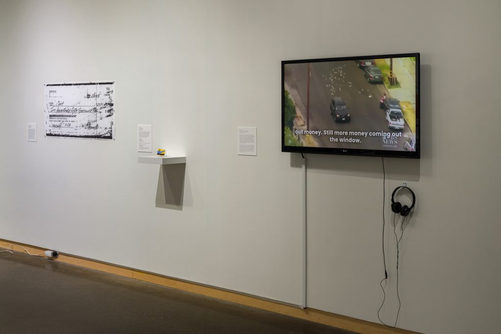 In a brightly lit gallery, a large printed check, small wall mounted case with small card, and mounted television and headphones, are spread across the wall. On the television, the scene includes money being thrown from a car.