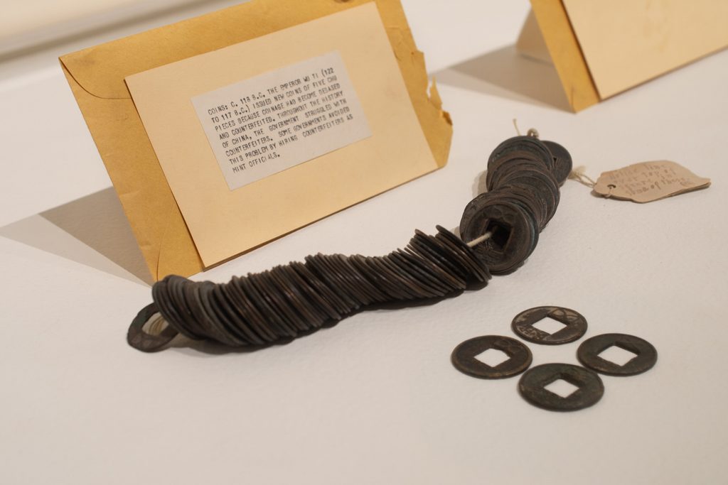 Loose and strung together ancient coins sit on top of a white background. A typed description on a yellow envelope sits in the background.
