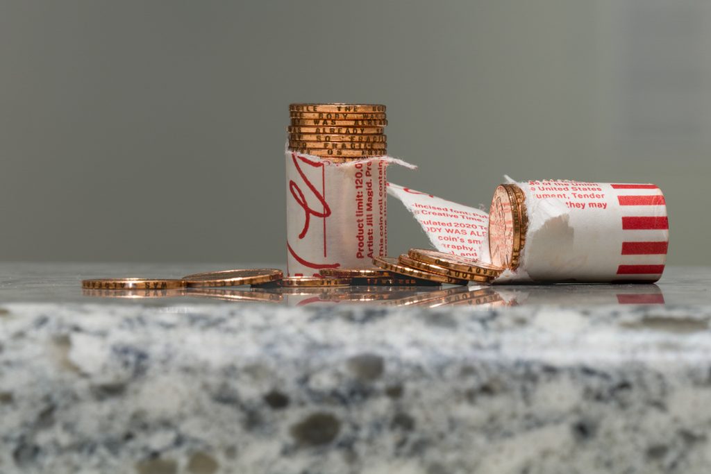 Alt text: A broken white and red paper roll of shiny pennies sit on top of a piece of black and white marble. On the edges of each penny reads “the body was already so fragile”.