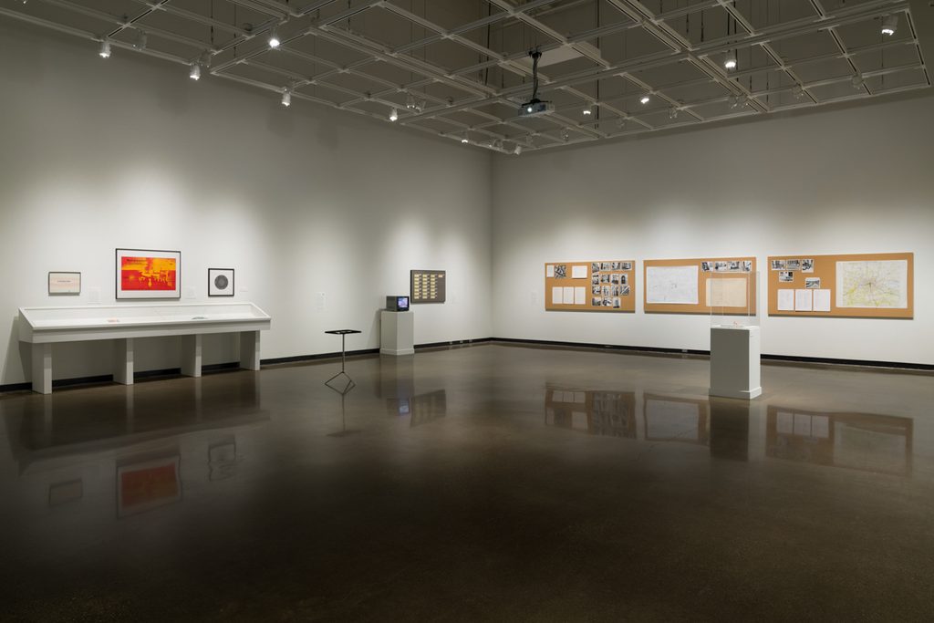 In a brightly lit gallery with concrete floors and high ceilings three large cork boards with a variety of maps, lists, and photographs are mounted to the right wall. The adjoining wall includes mounted framed pictures, a wall case, felt dice table, and television sitting on a pedestal.