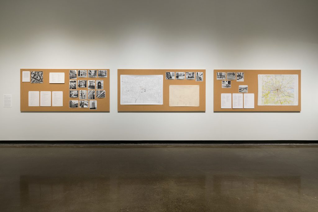 Three cork bulletin boards hang side by side in a brightly lit gallery. A variety of checklists, black and white pictures, and maps are hung with small silver and colorful pins.