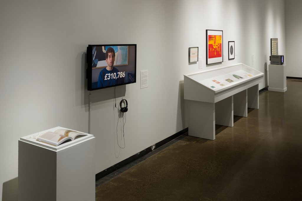 Along the wall of a brightly-lit galley sits an open book on top of a pedestal, a man on a television mounted on the wall, and three wall mounted artworks above a large case with smaller artworks.
