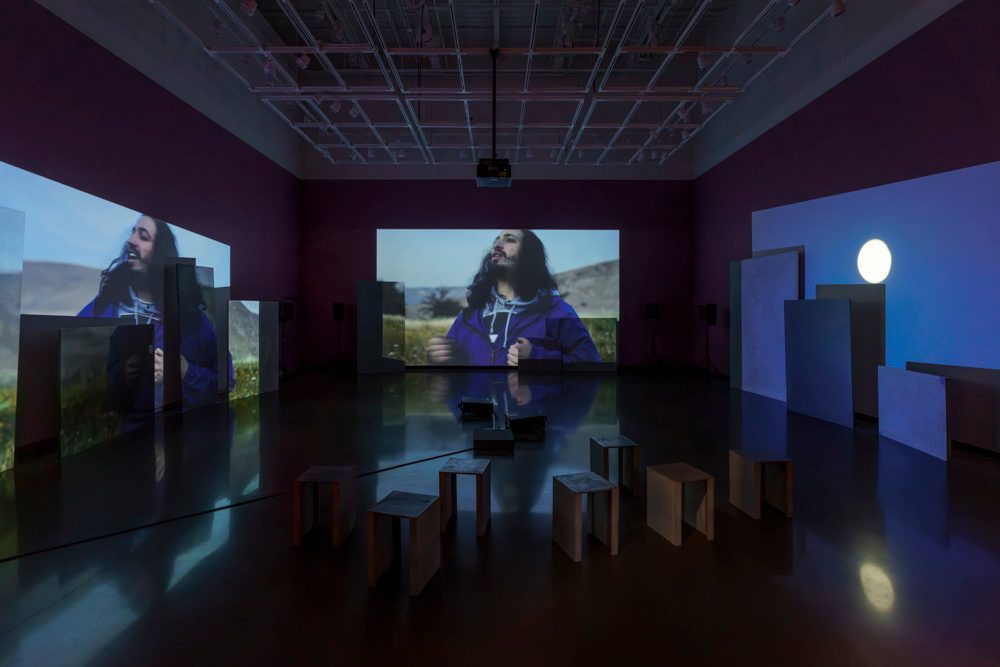 Three channels of video are projected onto uneven surfaces of large overlapping rectangular panels. The left and middle projection are different angles of a person dancing. The right projection is a full moon. In the foreground sit short, metal benches.