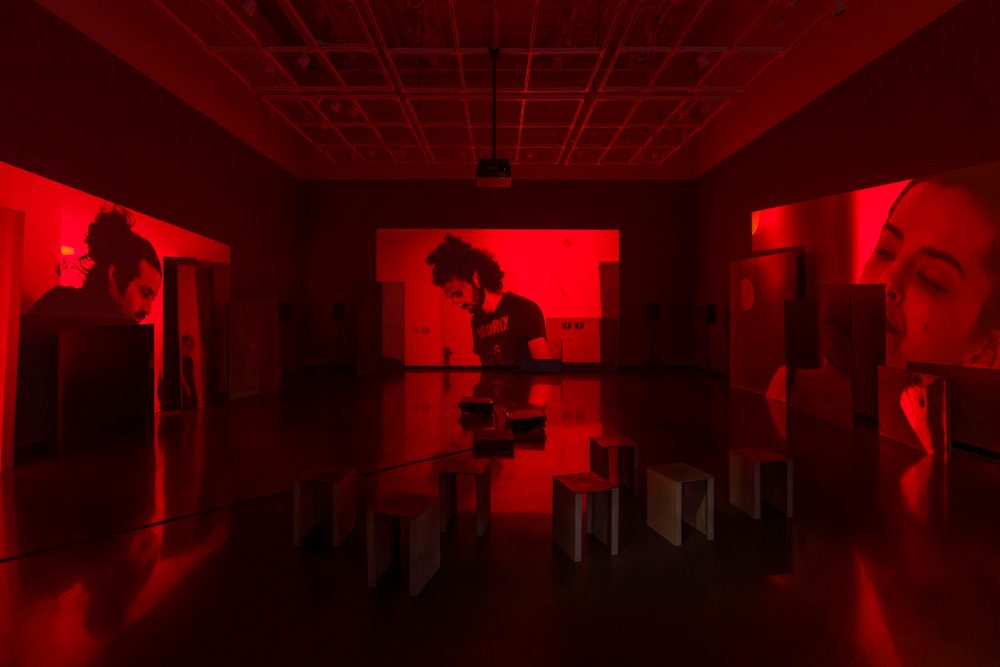 Three channels of video are projected onto uneven surfaces of large overlapping rectangular panels. The projections show three different shots of dancers in a red monochromatic lighting. In the foreground sit short, metal benches.