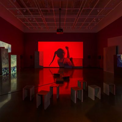 Three channels of video are projected onto uneven surfaces of large overlapping rectangular panels. The outside projections show inverted blue images. The middle projection shows a person in red, monochromatic lighting, dancing with a long swinging ponytail and their arms in the air. In the foreground sit short, metal benches.