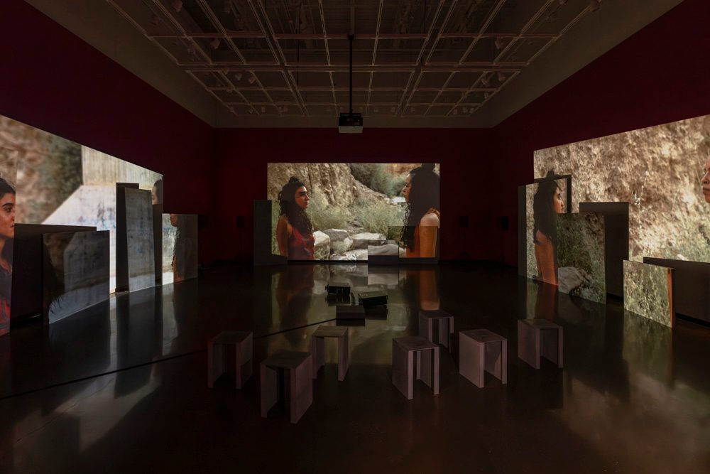 Three channels of video are projected onto uneven surfaces of large overlapping rectangular panels. Each projection shows a different angle of two people dancing and looking at each other with rocks in the background. In the foreground sit short, metal benches.