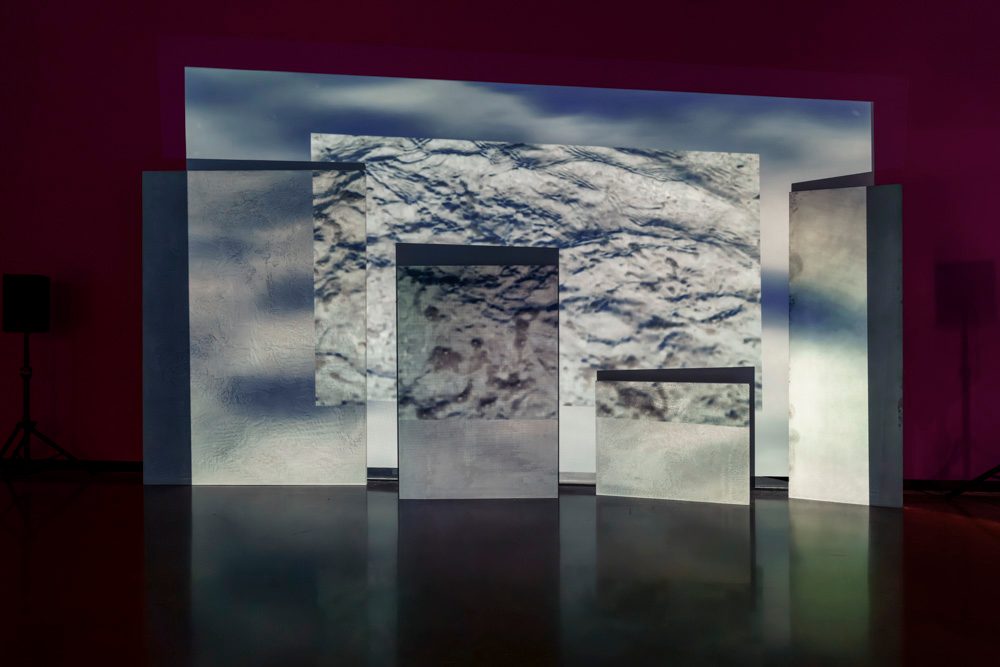A projection of a blue and white rough surface is projected across a wall and on various sized staggered, rectangular panels.