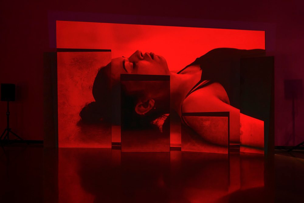 A projection of a person laying down with their eyes closed, in red light, is projected across a wall and on various sized staggered, rectangular panels.