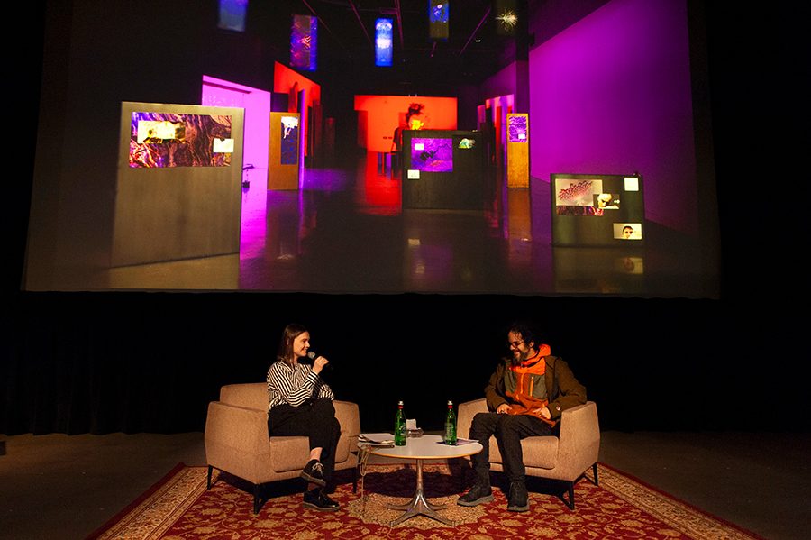 An interview of two people in a cozy setting with cushioned chairs, a table, and rug. Behind the speakers is a large projection of an art gallery.