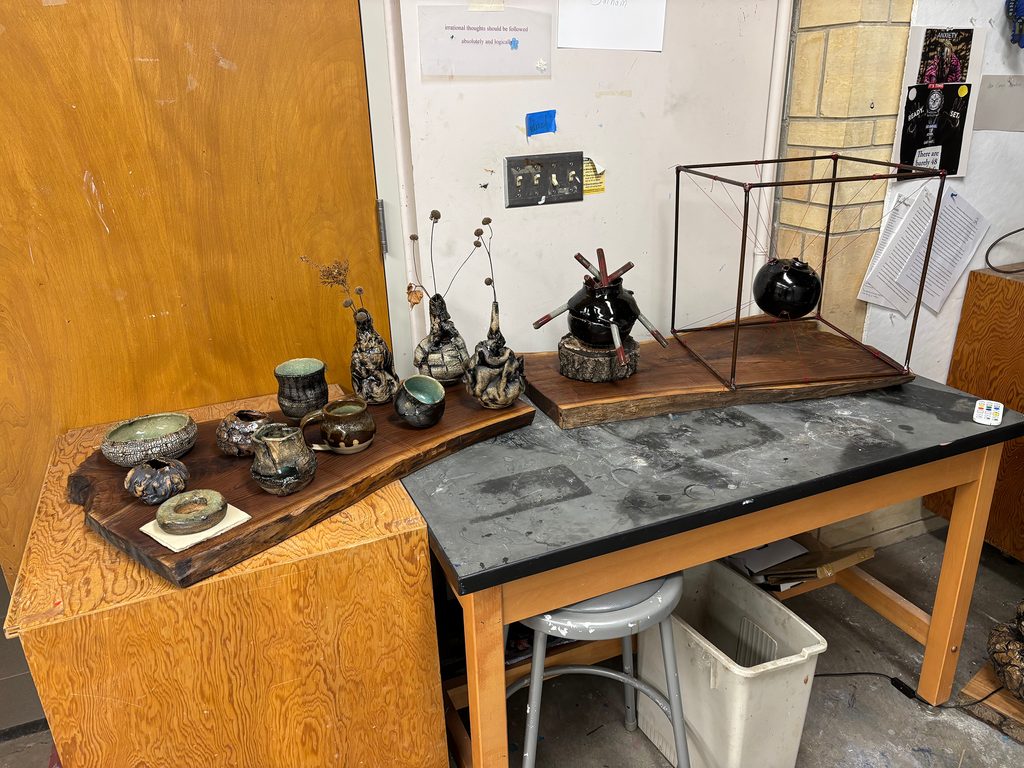 A series of ceramic sculptures on a workbench in a studio, additional elements are crafted from stoneware, glaze, steel round rod, and red string.