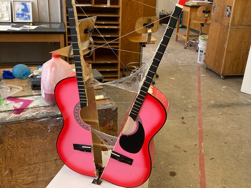 An abstract sculpture constructed from a pink acoustic guitar split in half, with the halves connected at a distance by silicone adhesive and steel wire