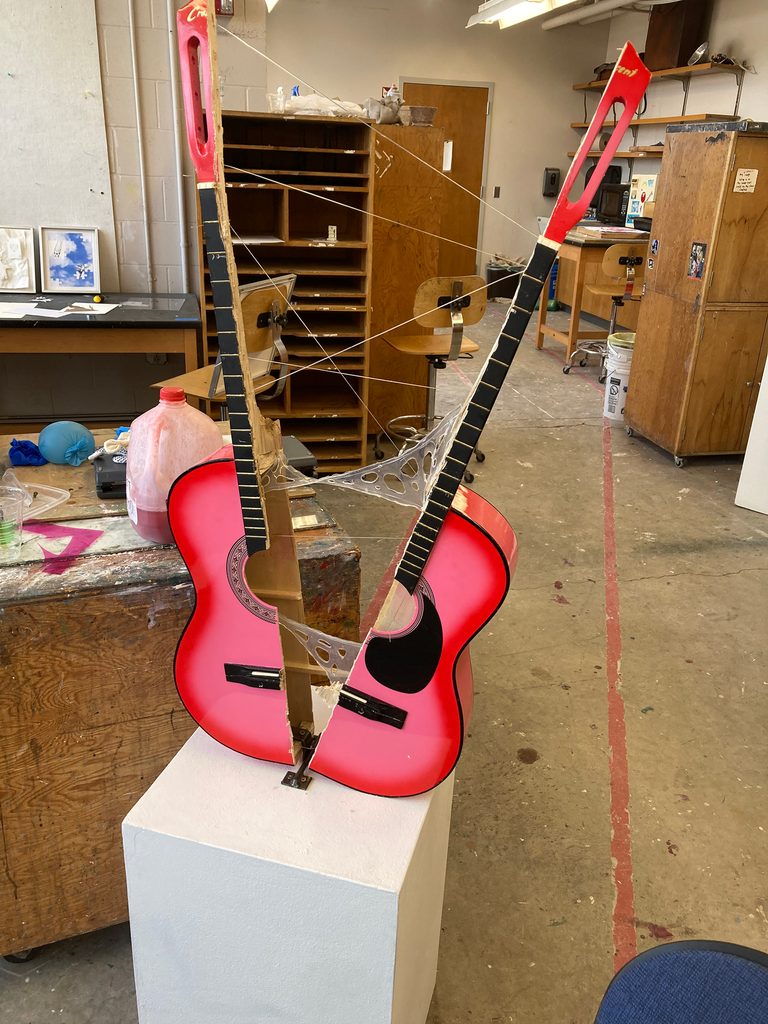 An abstract sculpture constructed from a pink acoustic guitar split in half, with the halves connected at a distance by silicone adhesive and steel wire