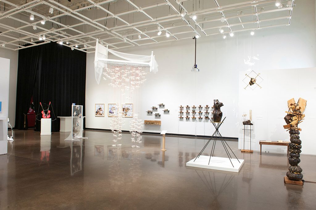 Many different types of artworks are displayed throughout a brightly-lit art gallery with white walls. A white bed with stringed ceramic horses hangs from the ceiling grids. Vertical woodland ceramics are to the right. A large black curtain hangs from the ceiling in the back right. pieces.
