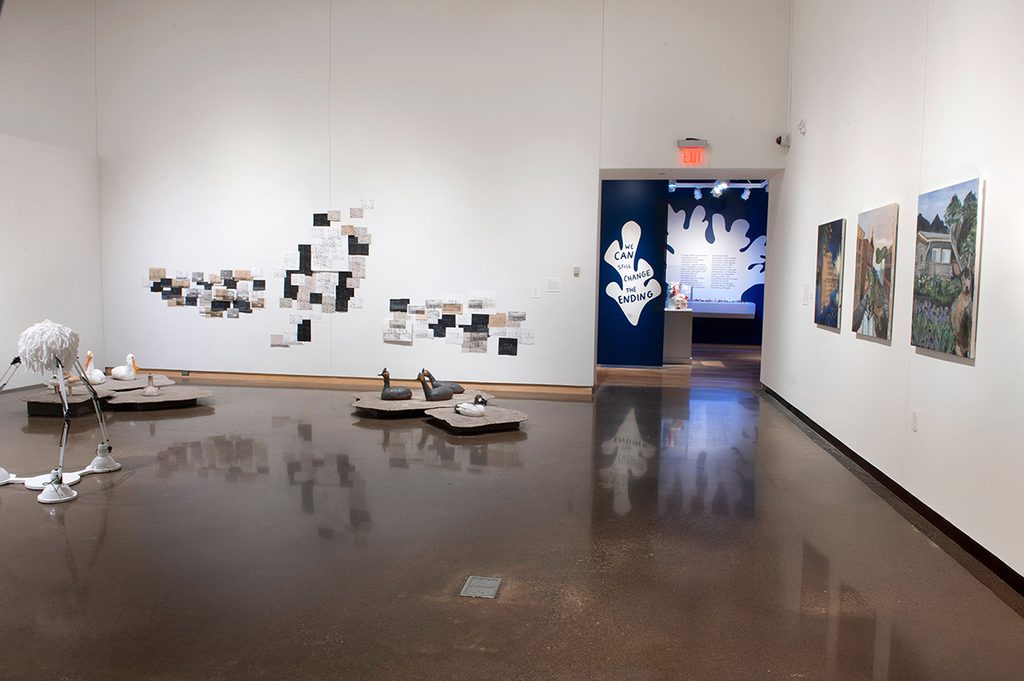 In a brightly lit gallery, with white walls, artwork hangs on a wall, and freestanding pieces are scattered along the floor. To the right is a entrance to another gallery with dark blue and white walls.