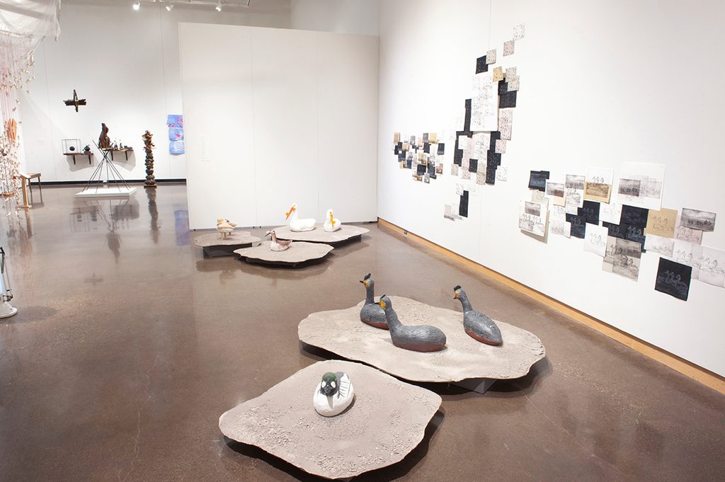 In a brightly lit gallery with white walls, ceramic birds sit on platforms with layered artwork prints mounted on on the right wall.