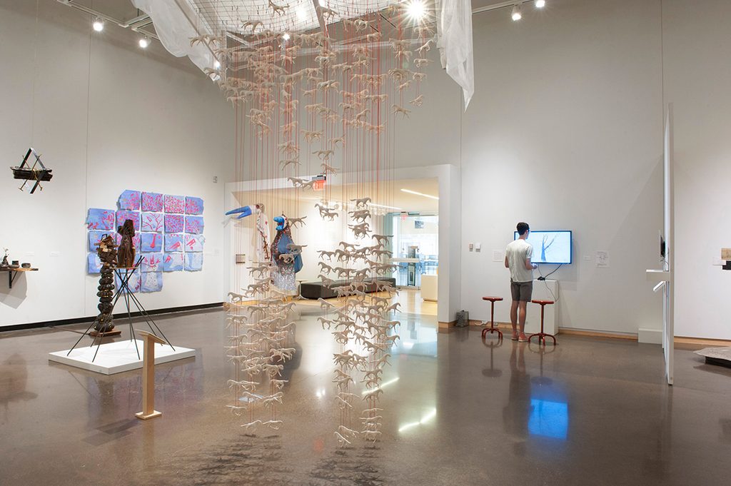 A group of artists display their work throughout a brightly-lit art gallery with white walls. A man stands in front of a monitor mounted to the wall. A bed frame hangs from the ceiling and has horses handing from red strings. On the left wall is collection of red, blue, and white paper in horizontal and vertical rows.