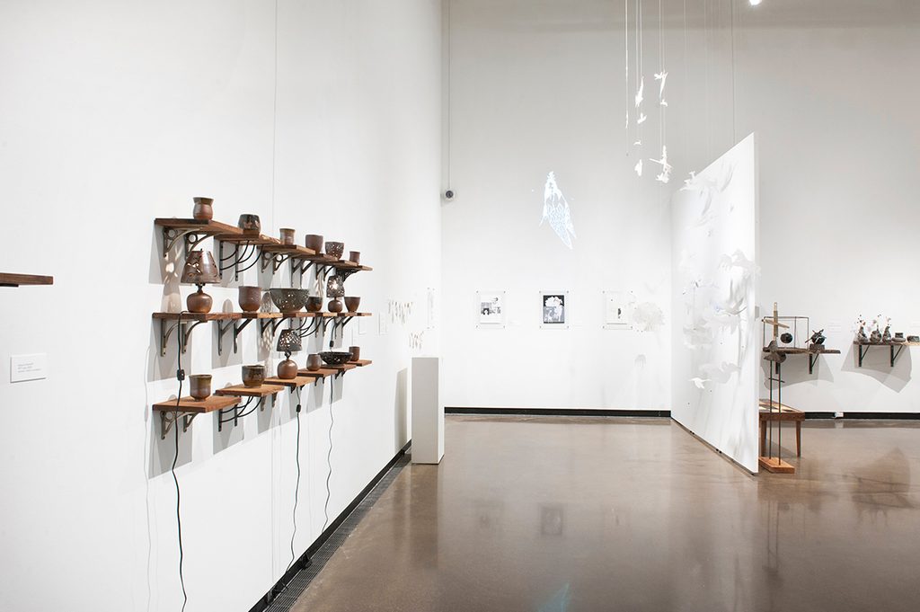 On the left are rows of ceramics sitting on small shelves, mounted to a wall in a gallery with brightly lit, white walls. Directly ahead a artist hung paper from the ceiling, mounted black and white prints on the wall, and an image was projected on the top of the wall.