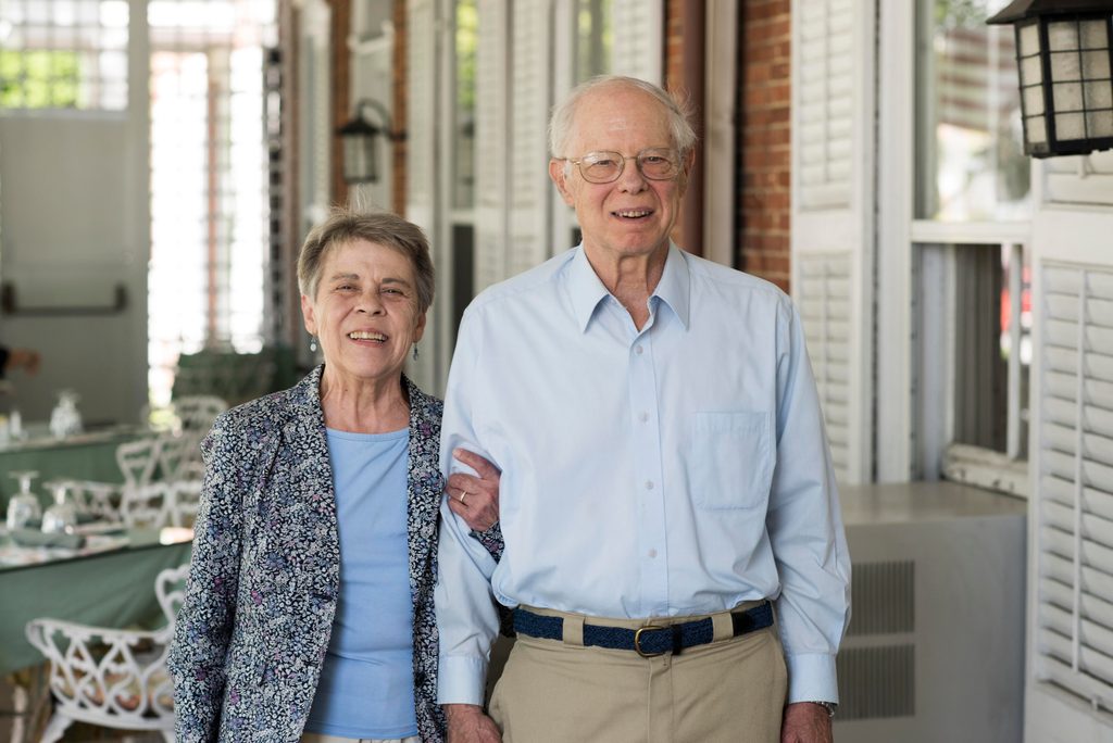 An elderly couple pose arm-in-arm, smiling