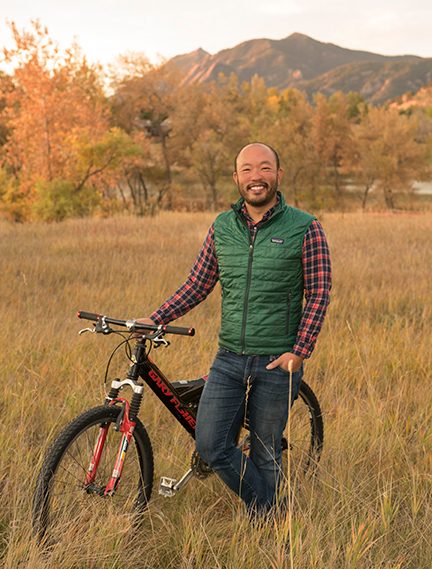 A man stands in a meadow beside a bicycle with a mountain in the background