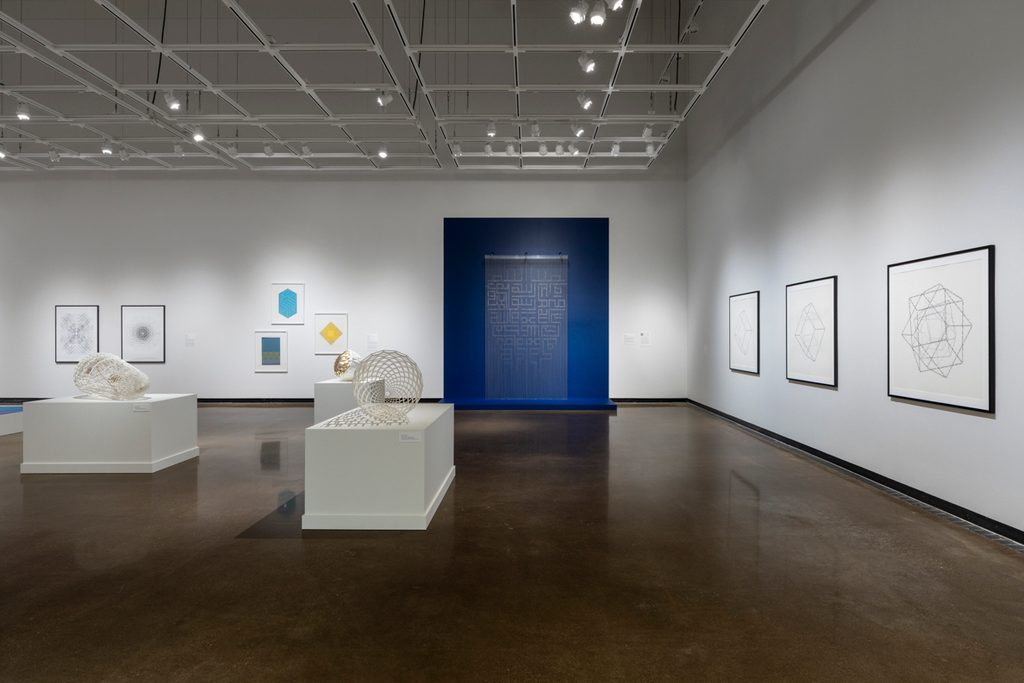 In an art gallery with white walls and high ceiling, geometric sculptures sit on white pedestals and framed drawings are hung on the wall. An art piece with rows of chains and beads is mounted on the back wall, framed by a painted blue rectangle.