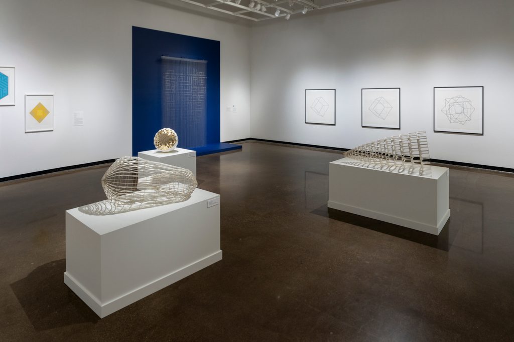 n an art gallery with white walls and high ceiling, geometric sculptures sit on white pedestals, and framed drawings are hung on the wall. An art piece with rows of chains and beads is mounted on the back wall, framed by a painted blue rectangle.