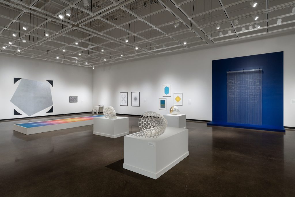 In an art gallery with white walls and high ceiling with grids, geometric sculptures sit on white pedestals, and framed drawings are hung on the wall. A beaded art piece is mounted on the back wall, framed by a navy rectangle painted on the wall. Colorful, framed artworks are also visible on the walls.