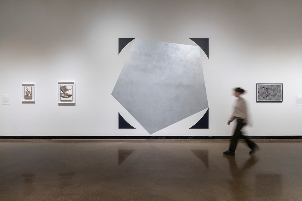 A person walks through a gallery with white walls. Artworks hang on the wall. In the middle are painted black and silver shapes.