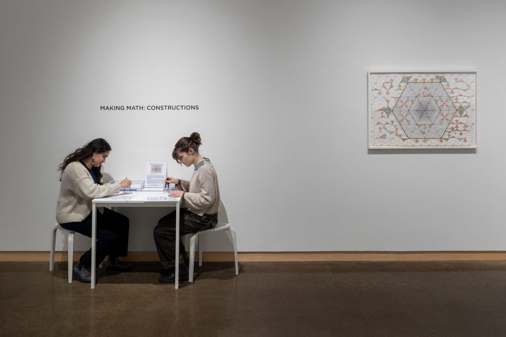 Two people sit across from each other at a small table in a museum gallery. Behind the reads “Making Math: Constructions.” A framed geometric artwork hangs to the right.