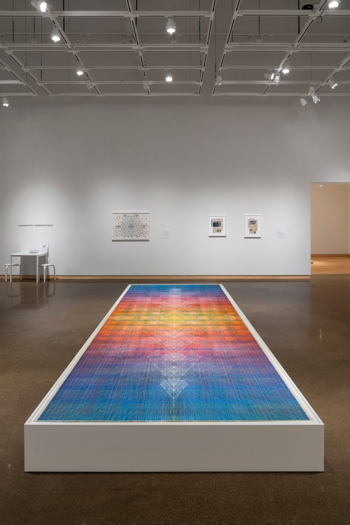A large, colorful rectangular artwork is displayed on a low platform in the center of the room. On the back wall hang framed geometric artworks.