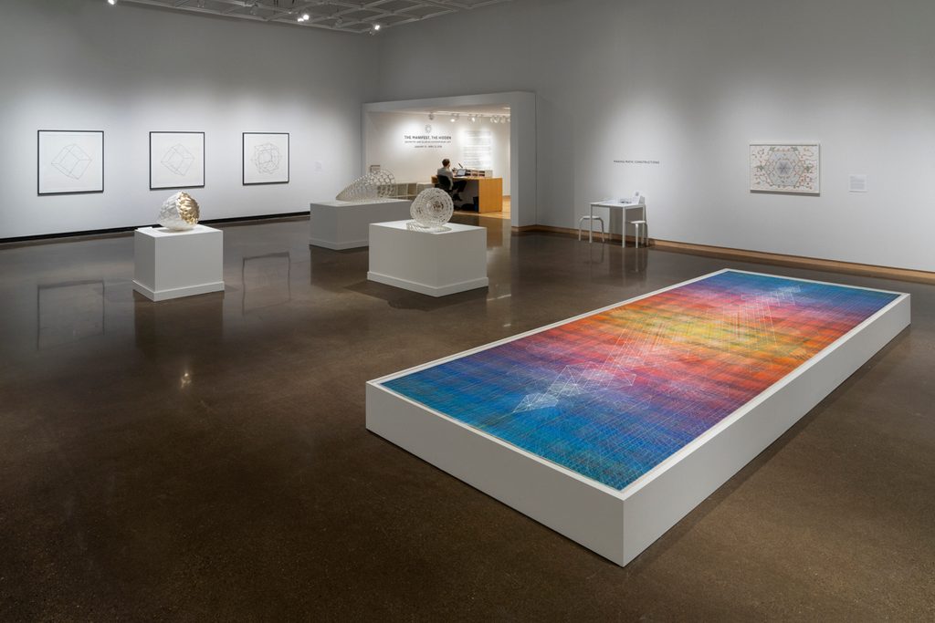 A large, colorful rectangular artwork is displayed on a low platform in the center of the room. Geometric pieces sit on white pedestals. On the back wall hang framed geometric artworks.