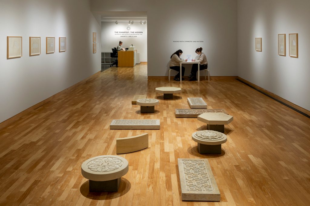 Stones slabs of different shapes, with geometric designs carved onto their faces, are scattered throughout the middle of a small art gallery with white walls and wooden floors. Framed works line the walls. In the background, two people sit face to face and are working on a table.