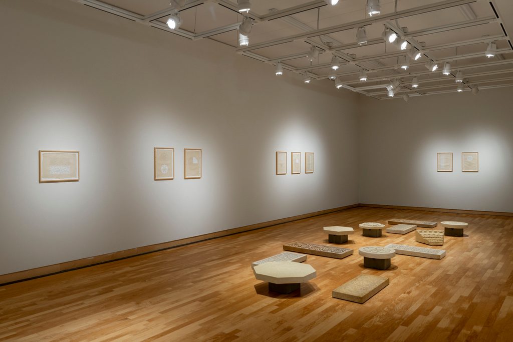 Stones slabs of different shapes, with geometric designs carved onto their faces, are scattered throughout the middle of a small art gallery with white walls and wooden floors. Framed works line the walls.