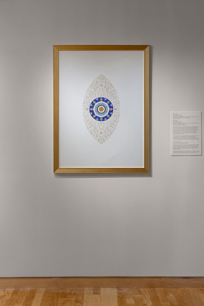 A large framed artwork with an oval shaped geometric design with gold and blue center is hung in a gallery with a white wall and wooden floor. A wall label is positioned to the right.
